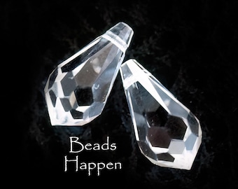 May include: Two clear, faceted crystal teardrop beads. The beads are transparent and reflect light, with a geometric design. The text "Beads Happen" is written in white script below the beads against a black background.