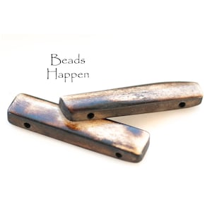 May include: Two brown and white rectangular wooden beads with holes drilled through them. The beads are stacked on top of each other. The text "Beads Happen" is printed above the beads.