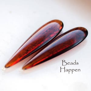 May include: Two teardrop-shaped glass beads in a deep red color with a marbled effect. Beads Happen is written in black text below the beads.