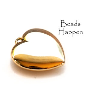 Puede incluir: Un charm dorado en forma de corazón con un acabado brillante. El charm tiene un pequeño bucle en la parte superior para sujetarlo a un collar o pulsera. El texto "Beads Happen" está escrito en negro encima del charm.