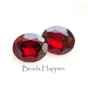 May include: Two faceted, oval-shaped, deep red gemstones. The gems are set against a white background. The text "Beads Happen" is written in a cursive font below the gemstones.