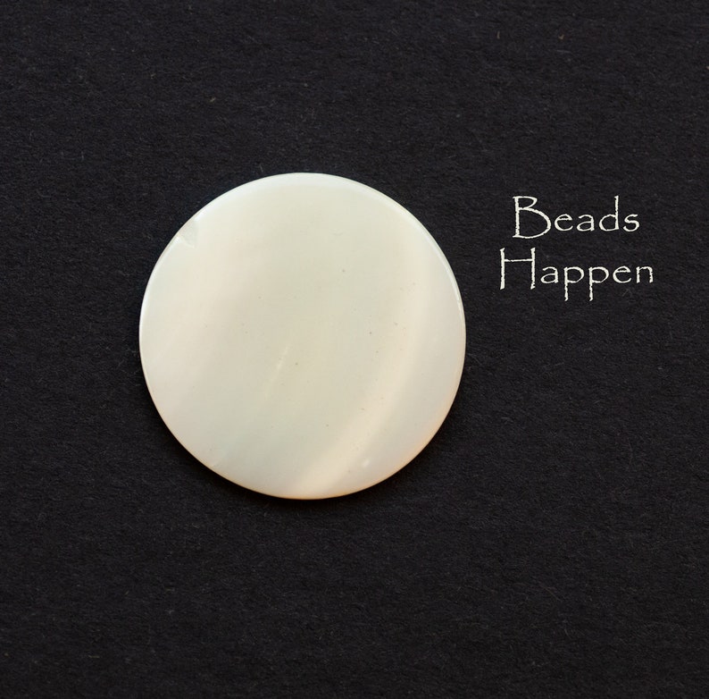May include: A white, round, smooth, and shiny bead. The bead is made of mother of pearl. The bead is on a black background. The text "Beads Happen" is written in white on the right side of the image.