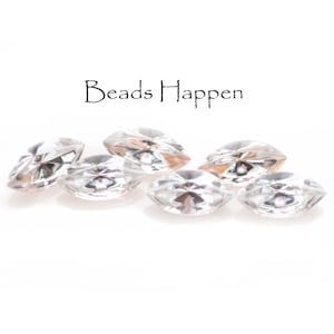 May include: Five clear faceted glass beads, each with a marquise shape, are scattered on a white surface. The text "Beads Happen" is visible above the beads.