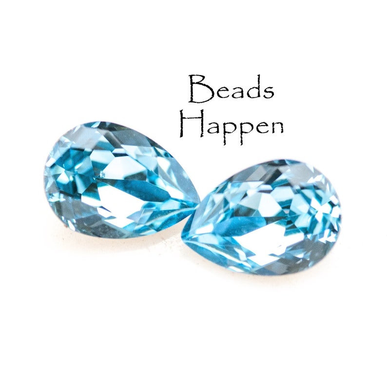 May include: Two pear-shaped, faceted, light blue gemstones are displayed against a white background. The gemstones are positioned close together, with the text "Beads Happen" above them.
