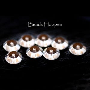 May include: Close-up of several clear, faceted beads with a dark brown center. The beads are arranged on a black surface. The text "Beads Happen" is in white script above the beads.