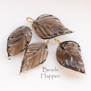 May include: Four smoky quartz leaf charms with gold-toned jump rings. The leaves have a detailed vein pattern and are translucent.