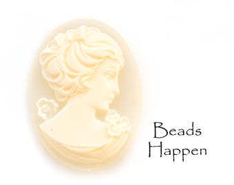 May include: Oval-shaped cameo pendant in a cream color, featuring a classical profile of a woman with detailed hair and floral accents. The text "Beads Happen" is printed in black.