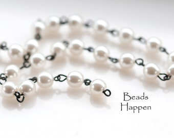 Op de afbeelding: Een ketting van witte imitatieparels verbonden door zwart draad. De parels zijn rond en gelijkmatig verdeeld. De tekst "Beads Happen" staat rechtsonder.