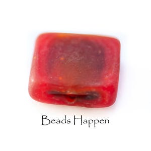 Puede incluir: Una cuenta roja de forma cuadrada con bordes redondeados. La cuenta es translúcida, con un centro más oscuro y bordes rojos más claros. El texto "Beads Happen" está escrito en escritura negra debajo.