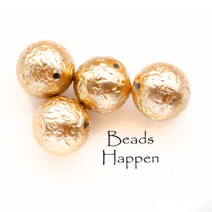 Puede incluir: Cuatro cuentas redondas de color dorado con una superficie texturizada. Las cuentas tienen un agujero en el centro para enhebrar. El texto "Beads Happen" está escrito debajo de las cuentas.