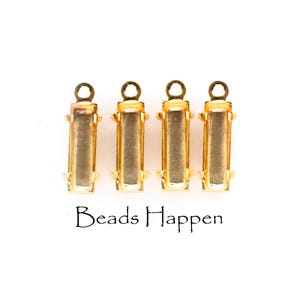 May include: Four gold-colored rectangular jewelry findings with a loop at the top and a rectangular opening. The text "Beads Happen" is printed in black script below the findings.