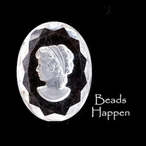May include: A clear glass oval bead with a raised cameo of a woman's profile. The bead has a faceted design and is set against a black background. The text "Beads Happen" is written below the bead.