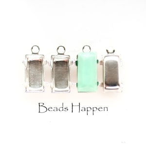 May include: Four rectangular jewelry components with silver-colored metal frames. One component features a light green, faceted gemstone. The text "Beads Happen" is printed in black below the components. These components are likely for jewelry making.