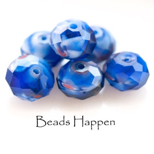 May include: A group of six blue and white glass beads with a faceted design. The beads have a swirl pattern and are arranged in a pile on a white surface. The text "Beads Happen" is printed below the beads.