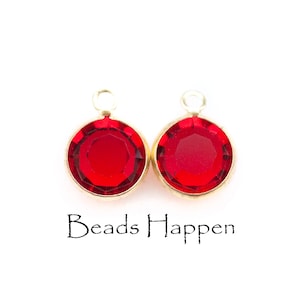 Puede incluir: Dos cuentas de cristal rojo con lazos de metal dorado. Las cuentas son redondas y tienen un diseño facetado. El texto "Beads Happen" está impreso debajo de las cuentas.