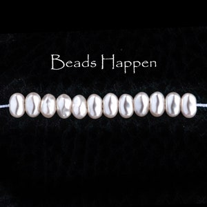 Puede incluir: Una hebra de cuentas blancas ovaladas con un ligero brillo. Las cuentas están enhebradas en un hilo blanco. El texto "Beads Happen" está escrito en blanco sobre las cuentas.