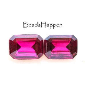 May include: Two vibrant pink emerald-cut gemstones, each with a smooth, polished surface. The stones are set against a white background.