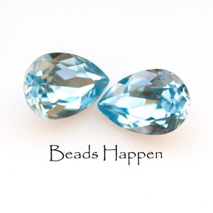May include: Two light blue, pear-shaped gemstones with multiple facets are displayed against a white background. The gemstones are positioned side by side. The text "Beads Happen" is written in black script below the gemstones.