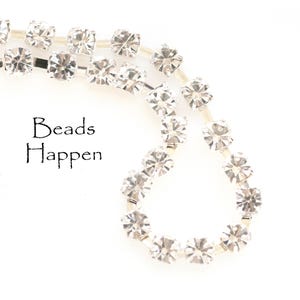 Puede incluir: Primer plano de un ribete de pedrería brillante con un diseño floral repetido. El ribete es plateado y transparente, con el texto "Beads Happen" en escritura negra.