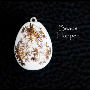 May include: White porcelain egg-shaped pendant with a gold floral design. The pendant has a loop at the top for hanging. The text "Beads Happen" is visible in the background.
