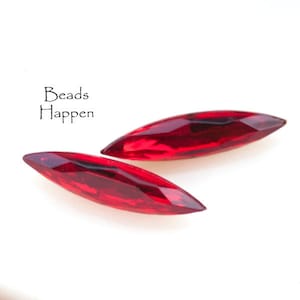 Puede incluir: Dos cuentas de cristal rojo oscuro, alargadas y facetadas. Las cuentas tienen forma de lágrima con extremos puntiagudos. La imagen incluye el texto "Beads Happen" en una fuente manuscrita.