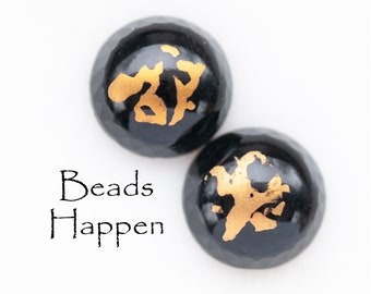 May include: Two black round beads with gold accents. The beads have a raised design on the surface. The text "Beads Happen" is printed below the beads.