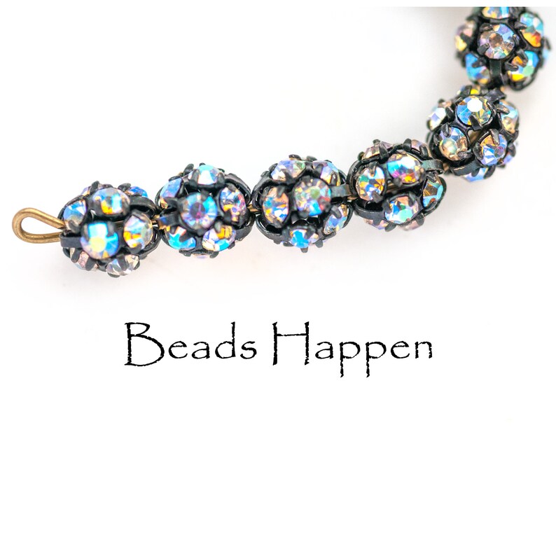 May include: A close-up of a beaded necklace with iridescent blue and black beads. The beads are round and have a faceted design. The necklace has a clasp at one end. The text "Beads Happen" is written below the necklace.