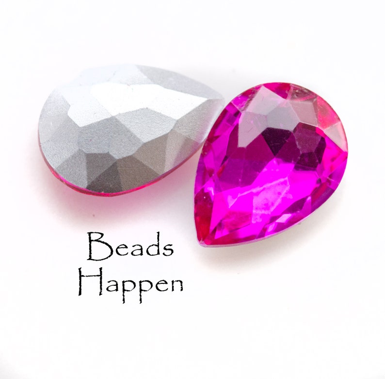 Puede incluir: Dos cuentas en forma de l&aacute;grima, una plateada y otra rosa brillante. Las cuentas son facetadas y tienen un acabado brillante. El texto "Beads Happen" est&aacute; impreso debajo de las cuentas.