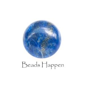 May include: A round, polished lapis lazuli gemstone with a deep blue color and flecks of gold. The gemstone is set against a white background. The text "Beads Happen" is below the gemstone.