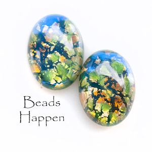 May include: Two oval-shaped glass beads with a blue, green, and gold iridescent finish. The beads have a speckled pattern and are arranged side-by-side on a white surface. The text "Beads Happen" is printed below the beads.
