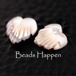 May include: Two white mother of pearl heart-shaped beads with a carved design. The beads are on a black background with the text "Beads Happen" written below them.