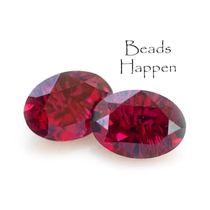May include: Two oval-cut, faceted gemstones in a deep red hue. The gems are displayed on a white background. The text "Beads Happen" is written in a cursive font above the gemstones.
