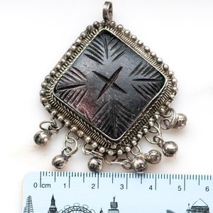 May include: A silver-toned pendant with a dark, carved central design. The pendant has a diamond shape with a beaded border and small bell charms hanging from the bottom. The carved design features a cross and linear patterns.