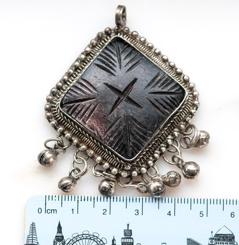 May include: A silver-toned pendant with a dark, carved central design. The pendant has a diamond shape with a beaded border and small bell charms hanging from the bottom. The carved design features a cross and linear patterns.