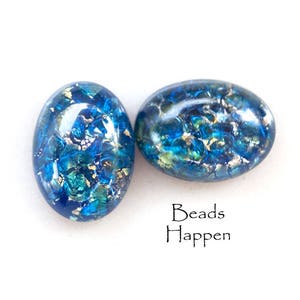 Op de afbeelding: Twee ovale kralen met een blauw, teal en goud gespikkeld ontwerp. De kralen hebben een glanzende afwerking tegen een witte achtergrond. De tekst "Beads Happen" staat eronder.