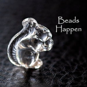 May include: A clear glass squirrel figurine with intricate details. The figurine is sitting on a black surface. The text "Beads Happen" is written in white above the figurine.