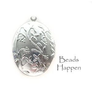 Op de afbeelding: Zilveren ovaal medaillon met een bloemmotief. Het medaillon heeft een scharnierende opening en een lus om een ​​ketting aan te bevestigen. De tekst "Beads Happen" is onder het medaillon gedrukt.