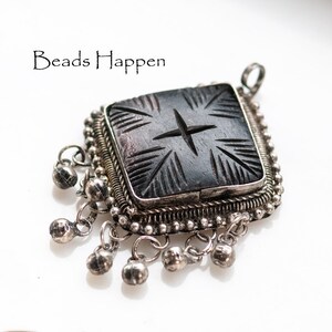 May include: A square, dark gray pendant with a carved starburst design, set in a silver frame. The pendant has small silver bells dangling from the bottom. The text "Beads Happen" is in the upper left corner.