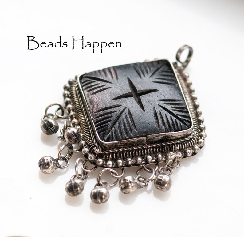 May include: A square, dark gray pendant with a carved starburst design, set in a silver frame. The pendant has small silver bells dangling from the bottom. The text "Beads Happen" is in the upper left corner.