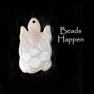 Puede incluir: Un colgante de tortuga de nácar blanco iridiscente. La tortuga tiene un caparazón detallado y es un diseño popular para la elaboración de joyas.