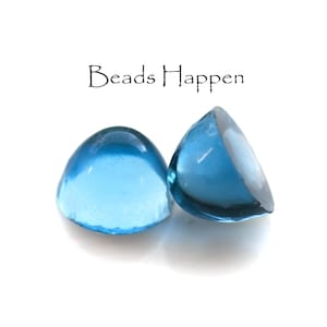 May include: Two blue glass cabochon beads, one is laying on its side and the other is upright. The text "Beads Happen" is written above the beads.