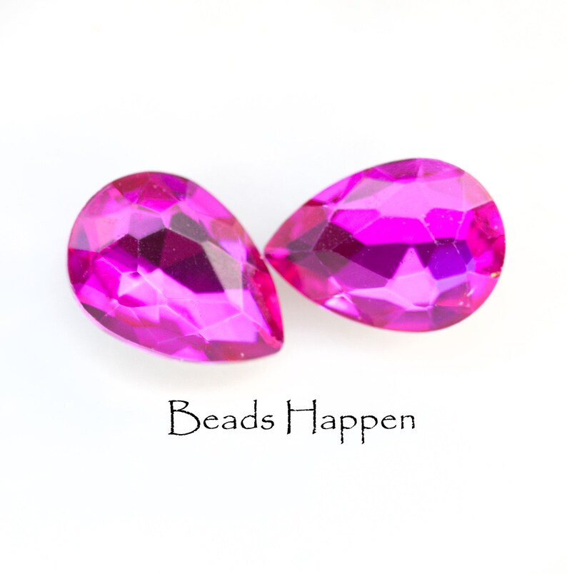 Puede incluir: Dos cuentas de vidrio de forma de l&aacute;grima, de color rosa nacarado. Las cuentas son facetadas y tienen un acabado brillante. Beads Happen est&aacute; escrito debajo de las cuentas.