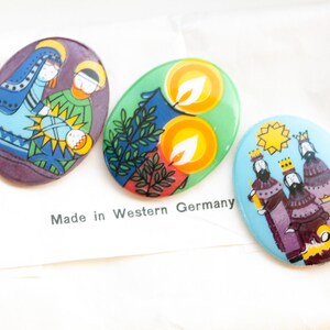 May include: Three oval-shaped Christmas ornaments with colorful illustrations. The first ornament depicts the nativity scene with Mary, Joseph, and baby Jesus. The second ornament features two yellow candles with green leaves. The third ornament shows the three wise men with a star above them. The ornaments are made in Western Germany.