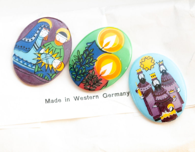 May include: Three oval-shaped Christmas ornaments with colorful illustrations. The first ornament depicts the nativity scene with Mary, Joseph, and baby Jesus. The second ornament features two yellow candles with green leaves. The third ornament shows the three wise men with a star above them. The ornaments are made in Western Germany.