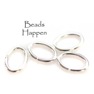 May include: Four silver-colored oval jump rings are scattered on a white background. The jump rings are open and ready to be used in jewelry making. The text "Beads Happen" is in black script.