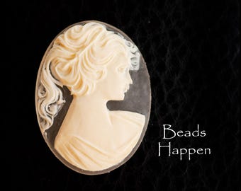 May include: Oval cameo pendant featuring a profile of a woman with flowing hair and draped clothing, in a cream color. The cameo is set against a clear background. The text "Beads Happen" is in white script on the right side.