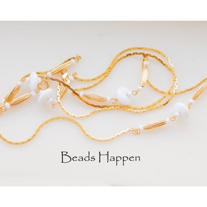 Pode incluir: Um colar de corrente dourada com contas brancas e detalhes dourados. O colar é em camadas e tem um design delicado. O texto "Beads Happen" é visível na imagem.