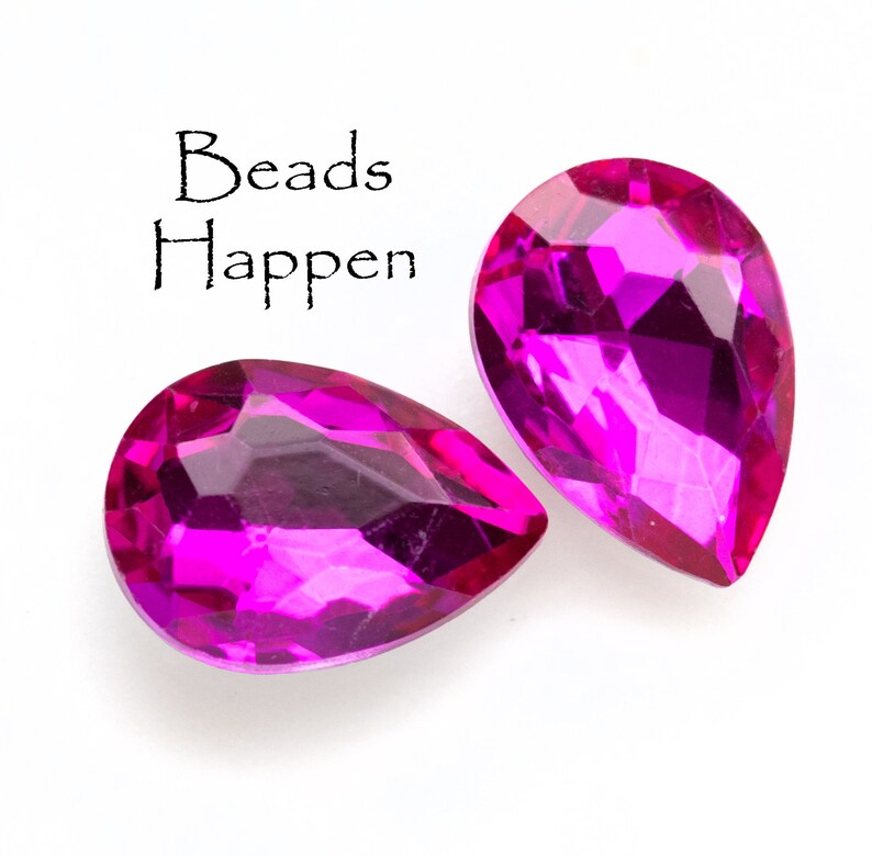 Puede incluir: Dos cuentas de cristal de color rosa brillante en forma de l&aacute;grima. Las cuentas son facetadas y tienen un acabado brillante. El texto "Beads Happen" est&aacute; escrito en negro sobre las cuentas.