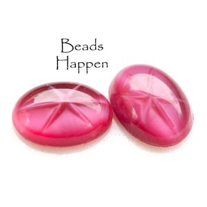 May include: Two pink oval-shaped cabochon beads with a star-shaped pattern. The beads are smooth and shiny.
