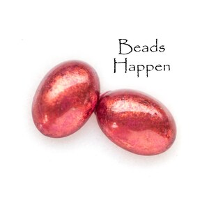 May include: Two red glass beads with a metallic sheen. The beads are oval shaped and have a smooth surface. The text "Beads Happen" is written above the beads.